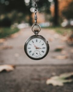 A vintage pocket watch hangs on a chain with a blurred outdoor path and scattered leaves in the background.