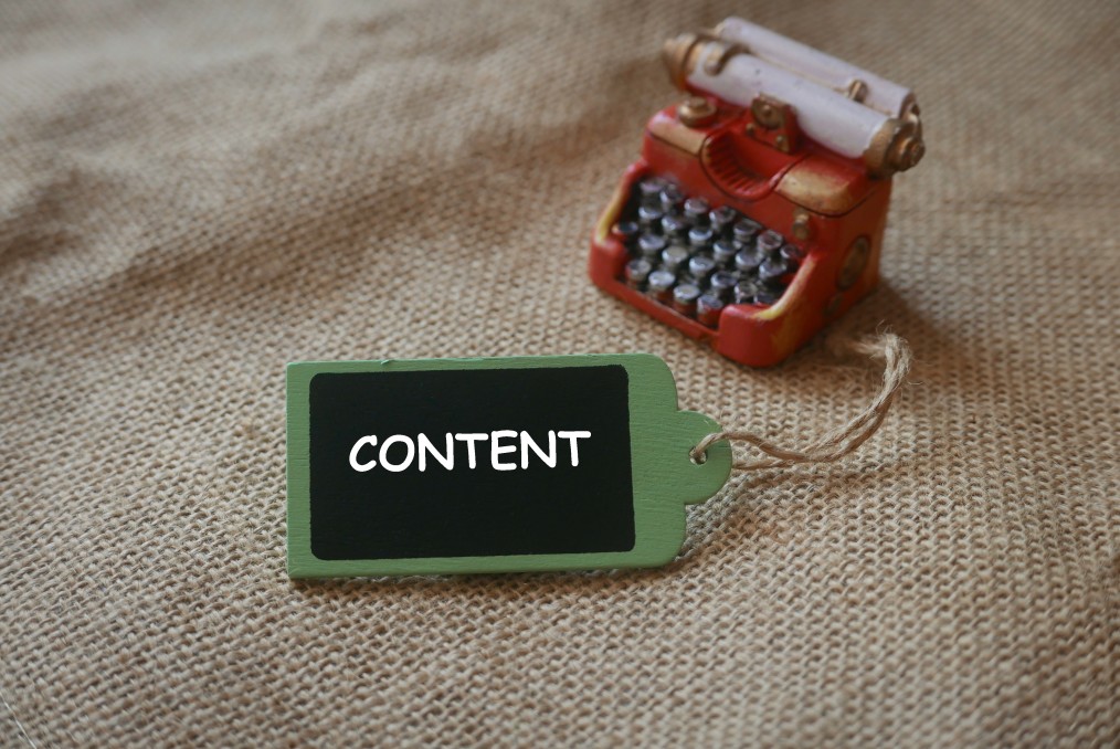 A small toy typewriter and a green tag with the word "CONTENT" written on it, placed on a burlap texture background.