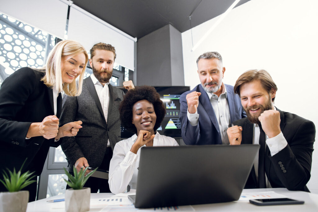 Five business professionals gather around a laptop in a modern office, smiling and making celebratory gestures, reflecting the collaborative spirit featured on our About Us page.
