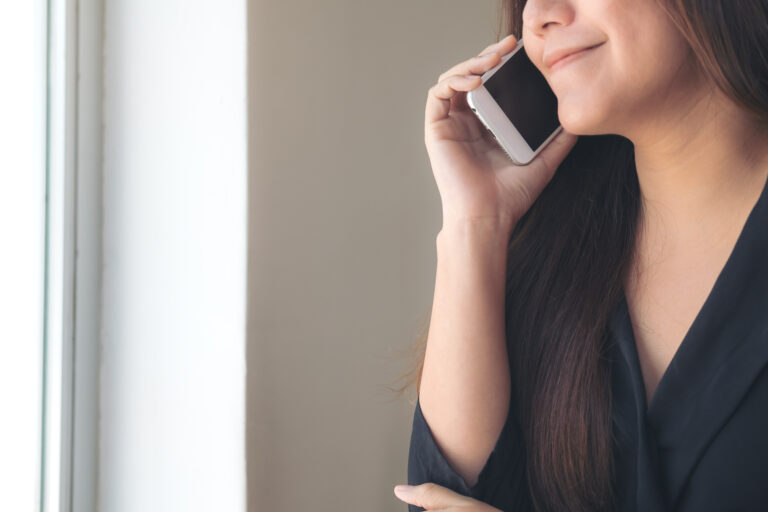 A woman with long dark hair is smiling while talking on a smartphone near a window, enjoying her first client call.