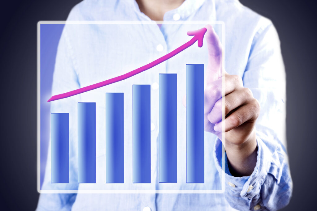 Person in a light blue shirt points at a virtual bar graph with ascending bars and a pink upward arrow, symbolizing business growth and the breakthrough needed to move beyond a business plateau.