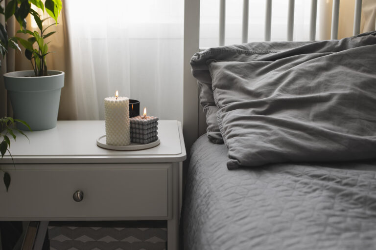 A white nightstand with two lit candles and a potted plant stands beside a neatly made bed with gray bedding in a softly lit bedroom, creating a peaceful space that encourages rest and productivity.