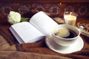 A cup of black coffee on a saucer sits beside an open book, a lit candle, and a white flower atop a brown blanket—the perfect setting to embrace rest strategy and restore work-life balance.
