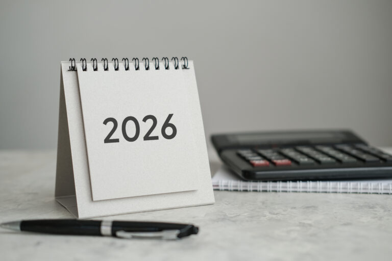 A desk calendar displaying "2026" next to a pen and a calculator on a spiral notebook, all placed on a light-colored surface—perfect for brand preparation 2026.