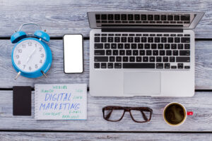 A laptop, smartphone, clock, coffee cup, glasses, business card, and a notepad with "Digital Marketing" terms are arranged on a wooden desk—ideal for sharing marketing lessons from 20 years experience in the digital marketing field.