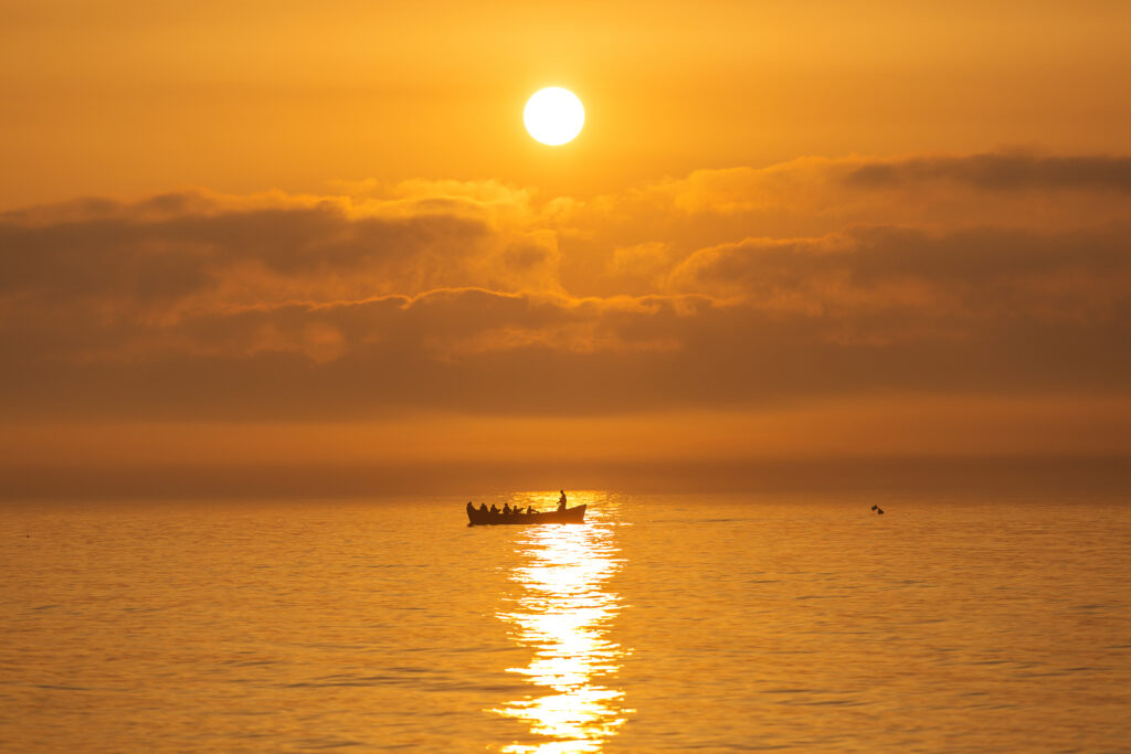 A group of people in a boat on calm water during a golden November sunset, with the sun low above the horizon and clouds in the sky.
