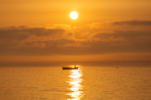 A group of people in a boat on calm water during a golden November sunset, with the sun low above the horizon and clouds in the sky.