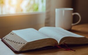 An open book with a red ribbon bookmark sits on a wooden table next to a white mug, sunlight streaming in—a peaceful setting for quiet reflection or planning your next strategy.