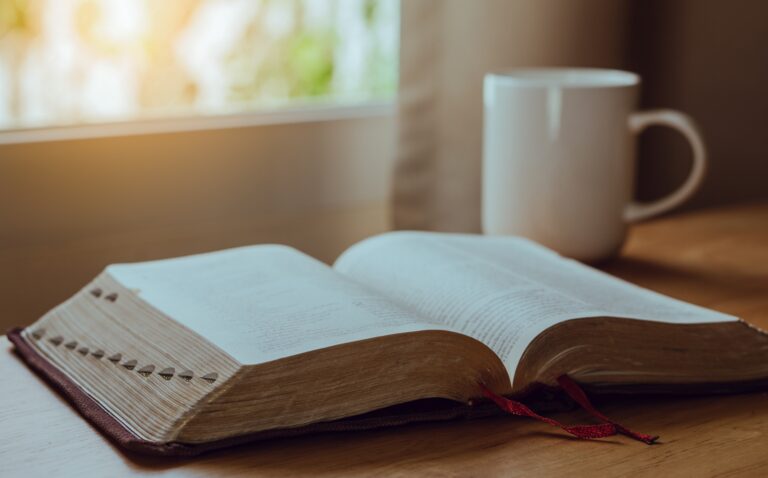 An open book with a red ribbon bookmark sits on a wooden table next to a white mug, sunlight streaming in—a peaceful setting for quiet reflection or planning your next strategy.