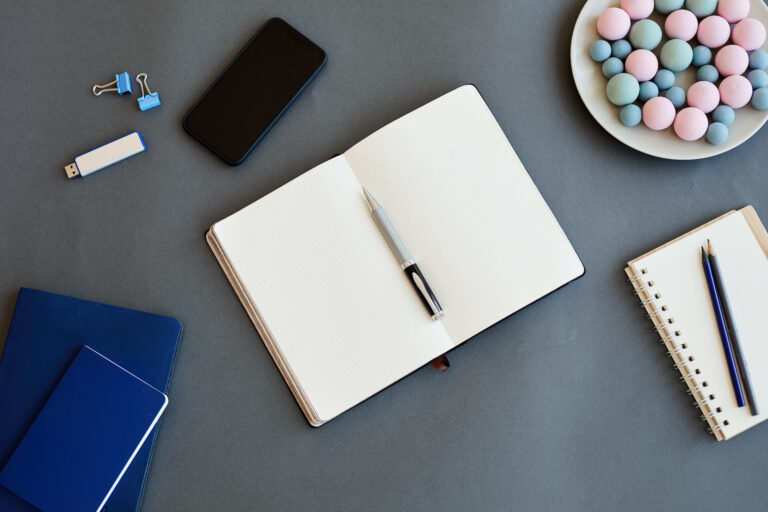 An open notebook with a pen, smartphone, USB stick, binder clips, and pastel candies on a plate sets the scene for clarity—not chaos—as you start the week. Two closed notebooks and another notepad with a pencil rest on a gray surface.