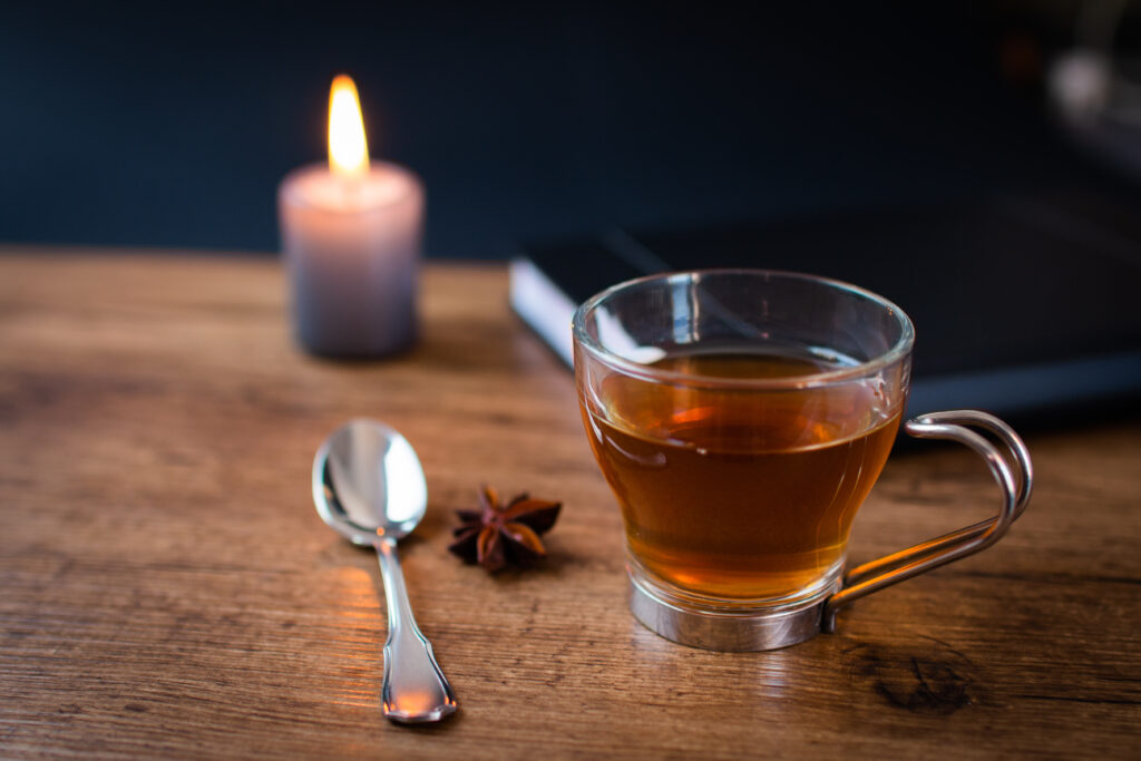 A glass cup of tea, a spoon, and a star anise rest on a wooden table—lit candle flickering nearby and closed notebook waiting—capturing a moment of quiet power before the rush.