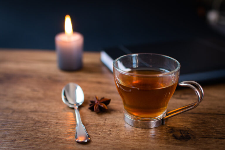 A glass cup of tea, a spoon, and a star anise rest on a wooden table—lit candle flickering nearby and closed notebook waiting—capturing a moment of quiet power before the rush.