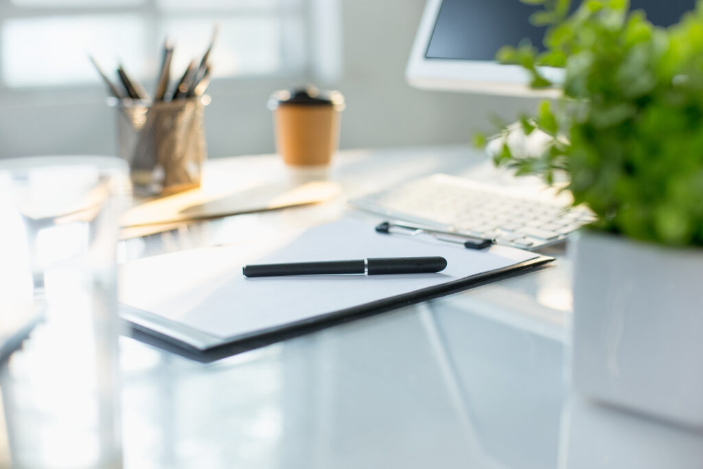 A black pen rests on a clipboard with paper on a desk, next to a computer keyboard, potted plant, and takeaway coffee cup—ready for client notes or lessons learned with patience.