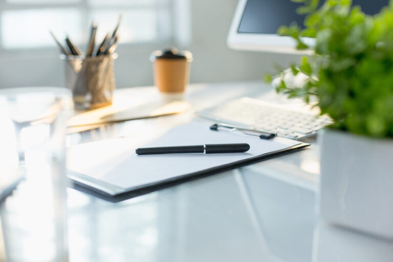 A black pen rests on a clipboard with paper on a desk, next to a computer keyboard, potted plant, and takeaway coffee cup—ready for client notes or lessons learned with patience.