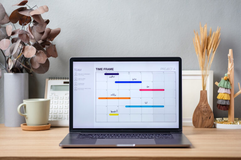 A laptop on a desk displays a project timeline chart, ideal for planning social media tasks. The desk also holds a calendar, cup, saucer, dried plants, and decorative items against a light wall to simplify your work environment.