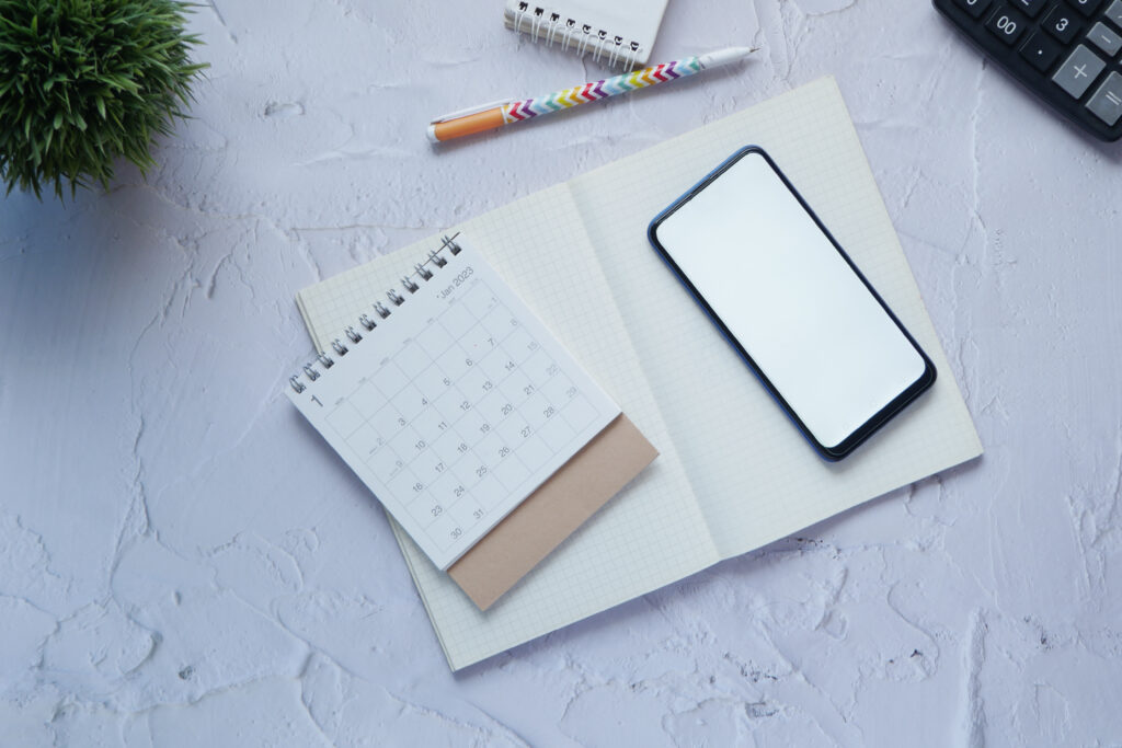 Open notebook with a blank phone screen, small calendar, pen, potted plant, and part of a calculator on a white textured surface—perfect for planning creativity in marketing or new marketing strategies.