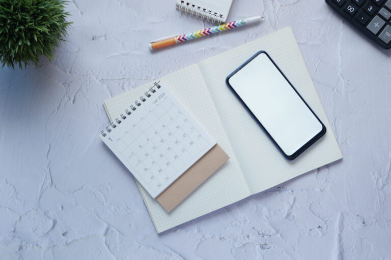 Open notebook with a blank phone screen, small calendar, pen, potted plant, and part of a calculator on a white textured surface—perfect for planning creativity in marketing or new marketing strategies.