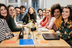 A group of twelve people sit around a wooden table, smiling toward the camera; laptops, notebooks, mugs, and plants are on the table—capturing the spirit of the People Behind the Progress.