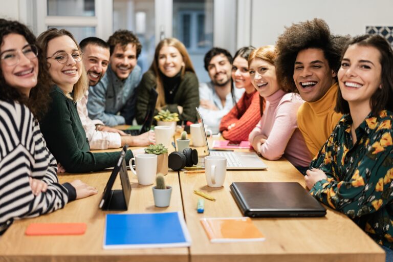 A group of twelve people sit around a wooden table, smiling toward the camera; laptops, notebooks, mugs, and plants are on the table—capturing the spirit of the People Behind the Progress.